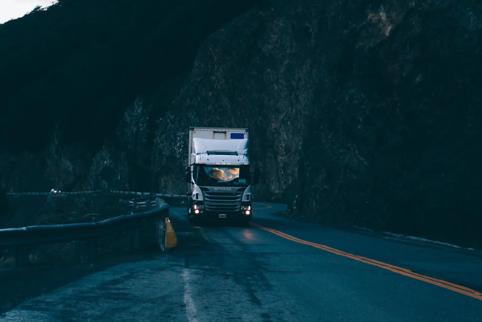Caminhão branco numa estrada de serra ao anoitecer, com os faróis acesos